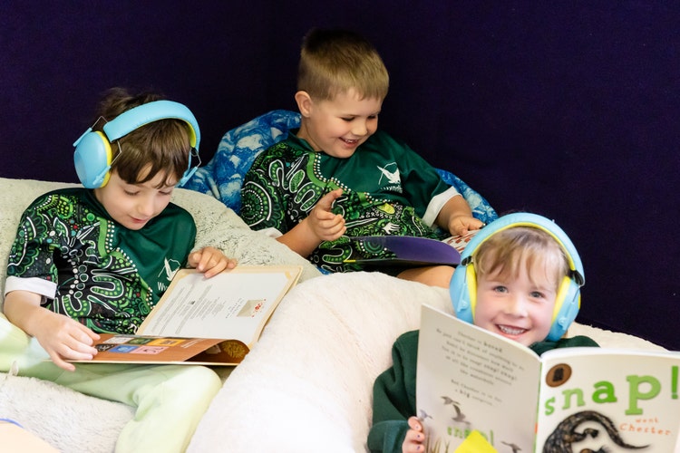 Three students sitting on beanbags reading books and wearing headphones.